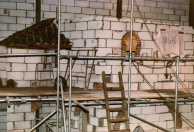 Armenwijk, 12-1984. Links boven de bedelaars in de Armenwijk zit dit, in basis, groene dakje verscholen. Later wordt dit zeer donker en dof gedecoeerd. Rechtsonder een boog als relief in de muur direct achter de bedelaars. De boog linksonderin is van een 'voordeur' links van de bedelaars. Rechts aan de wand hangt een bouwtekening van Ton van de Ven met daarop de detaillering van de scène.