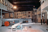 Opstaphal, 12-1985. We staan in de vaargeul van de opstaphal, waar werklieden op steigers bezig zijn met de afwerking en bekabeling van de ruimte. De bekabeling loopt tussen de dakspanten, die later worden afgedekt met plafondplaten. In de voorgrond een zaagtafel.