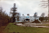 Exterieur, 04-1985. Het voorplein komt pas laat tijdens de bouw gereed - hier bestaat het nog grotendeels uit het een grasveld met daaromheen bosschages en bomen. Hier net buiten beeld staan de tijdelijke werkplaats en nissenhut en de restanten van de manege. Twee mannen werken aan de minaret.