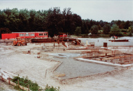 Exterieur, 05-1984. Precies een jaar geleden zijn de bouwtekeningen ingediend, enkele maanden geleden is gestart met de bouw. De oranje bouwketen zijn van hoofdaannemer Pepping uit Kaatsheuvel. We zien het bewapeningsijzer voor de fundering die de vaargeul gaat vormen. In de achtergrond de vijver, die pas later wordt uitgegraven tot aan de gevel. De Vonderplas is tijdens de bouw nog lang in gebruik als Roeivijver. Een gele boot is geparkeerd bij de rode truck, vlak voor het bouwhek dat de Vonderplas-route blokkeert.