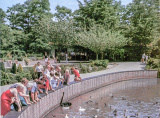 Bezoekers aan de rand van de <a href="/lemma/Siervijver" title="Siervijver">Siervijver</a> bij het <a href="/lemma/Caf%C3%A9-Restaurant" title="Café-Restaurant">Café-Restaurant</a>, waarvan nog een stukje terras te zien is. Tussen de bomen staan mensen bij <a href="/lemma/Station_Noord" title="Station Noord">Station Noord</a> van de <a href="/lemma/Efteling_Stoomtrein_Maatschappij" title="Efteling Stoomtrein Maatschappij">Efteling Stoomtrein Maatschappij</a>.