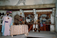 Tweede Marktscène, 02-1986. Close-up van de flessenverkoper, met nog opvallend weinig flessen, en de marktkoopman achter zijn toonbank. In de loop der jaren is de vulling van de toonbank aan verandering onderhevig en krijgt de plank met pantoffeltjes verschillende plekken in de scène. Wat opvalt is het grote gat in de voorgrond, de plek waardoor alle stoelen met nokkenschijven in de onderliggende kelders zijn geplaatst. Later wordt deze afgedekt en wordt het omliggende tegelpatroon doorgetrokken waardoor je het vrijwel niet kan zien.