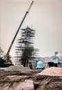 Exterieur, 06-1985. De koepel wordt op de minaret geplaatst, waarmee een van de laatste exterieur-bouwelementen gereed komt. In de voorgrond de typische kleine witte steentjes waarmee het voorplein bestraat wordt en die er de eerste twaalf jaar blijven liggen. De boom die hier nog naast de minaret staat wordt uiteindelijk verwijderd, vermoedelijk omdat die afbreuk doet aan het zicht op en/of de schaal van de imposante toren.