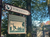 <a href="/lemma/Toilet" title="Toilet">Toilet</a>-wegwijzer bij <a href="/lemma/Carrouselpaleis" title="Carrouselpaleis">Carrouselpaleis</a>, ingang <a href="/lemma/Diorama" title="Diorama">Diorama</a>. Een jongeling rent naar de wc terwijl op de achtergrond een machinist een stoomtrein bedient.